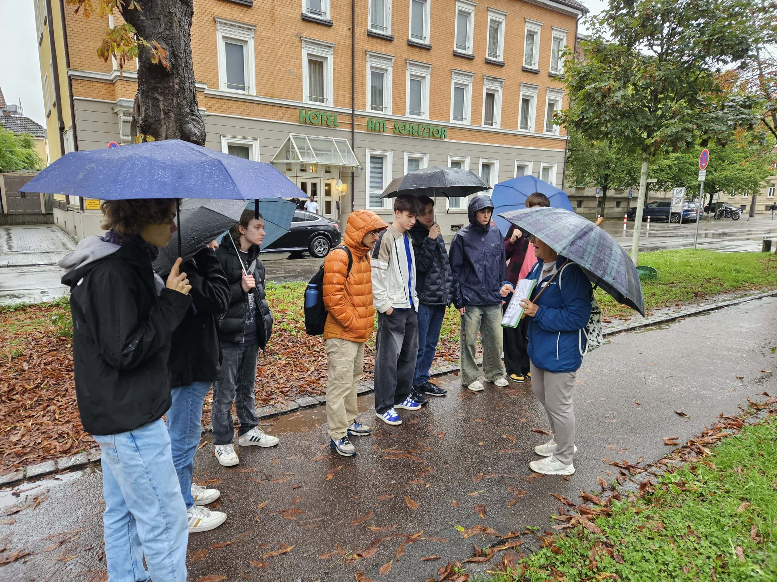 Gruppe von Menschen mit Regenschirmen im Regen, sprechen miteinander vor einem Hotel.