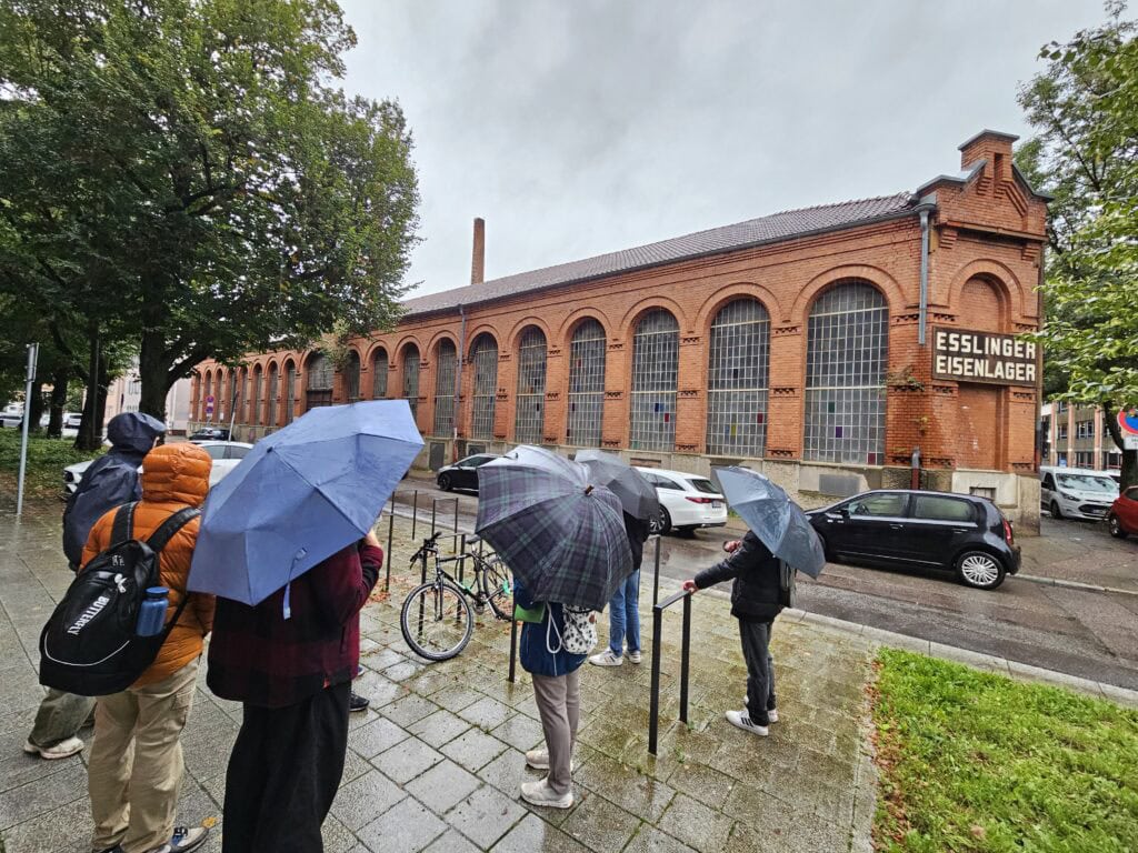 Menschen mit Regenschirmen vor einem roten Backsteingebäude mit der Aufschrift "Esslinger Eisenlager".