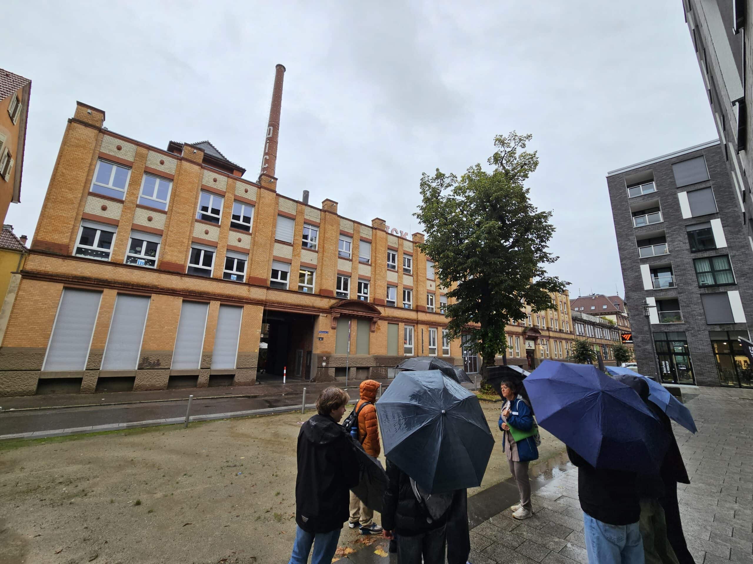 Gruppe von Menschen mit Regenschirmen, die vor einem historischen Gebäude stehen. Bewölkter Himmel.