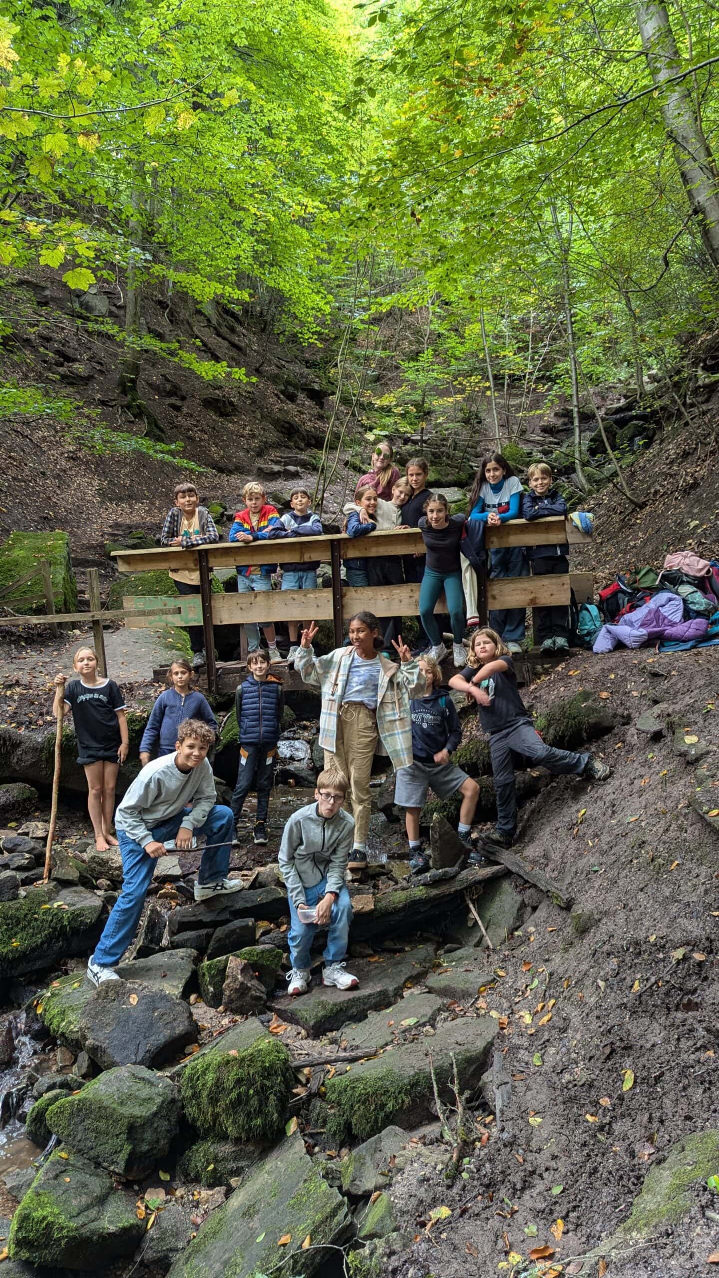 Eine Gruppe fröhlicher Kinder steht im Wald neben einem Bach, umgeben von Herbstblättern.