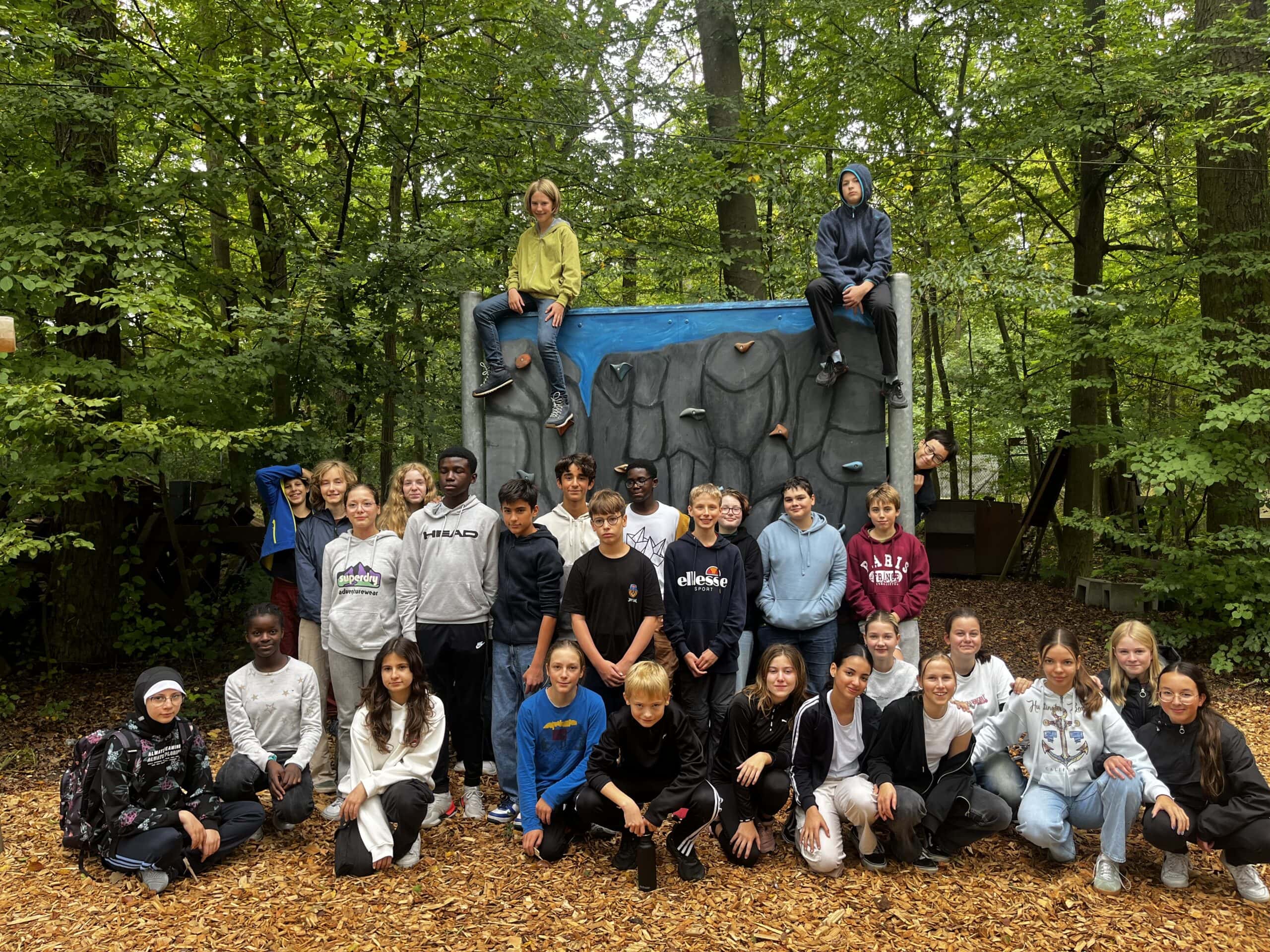 Eine Gruppe von Schülern posiert lachend vor einer Kletterwand im Wald; bunt gekleidet, Herbstlaub.