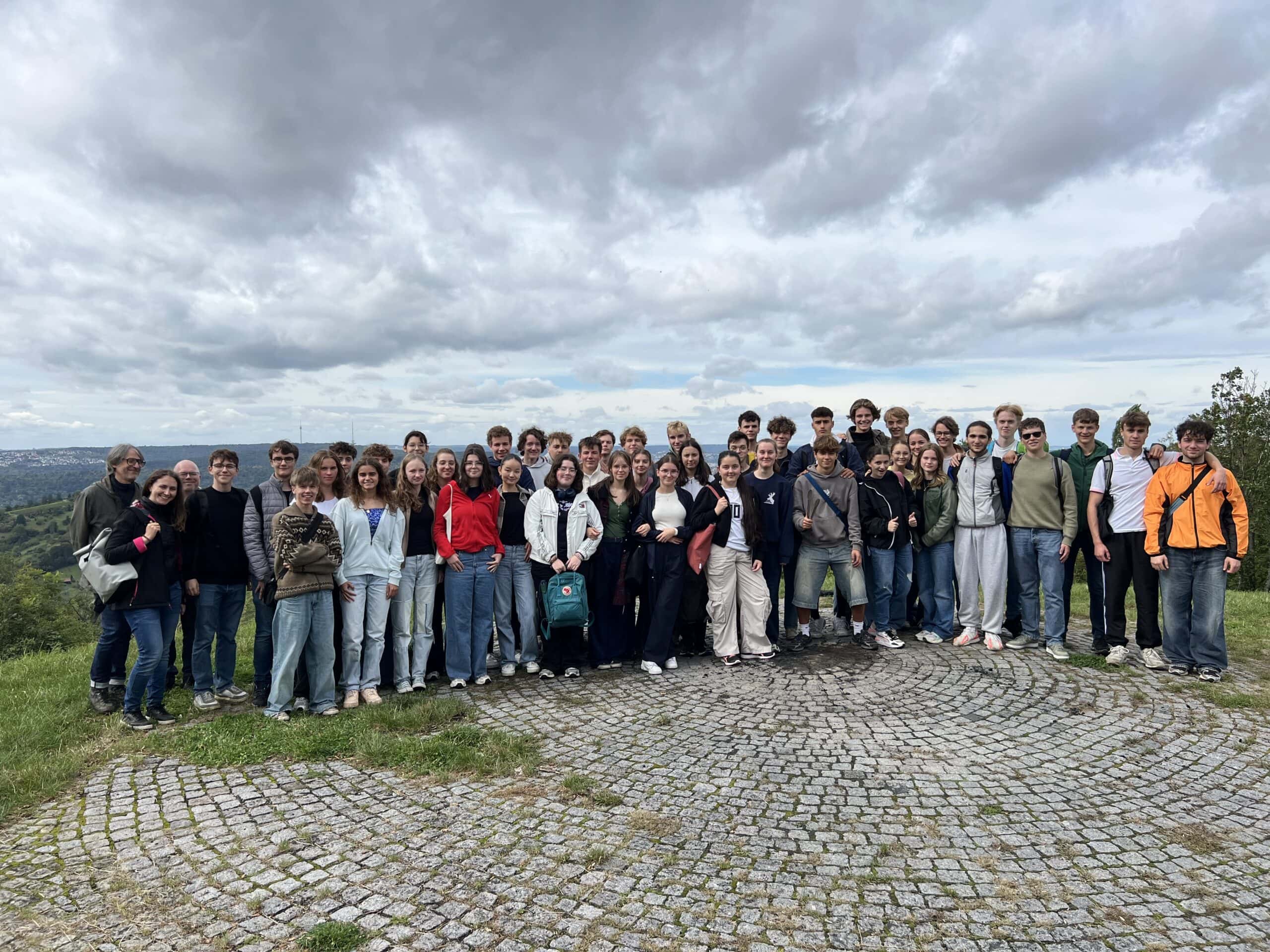 Eine Gruppe von Jugendlichen lächelt draußen, wolkiger Himmel, viele tragen Jeans.