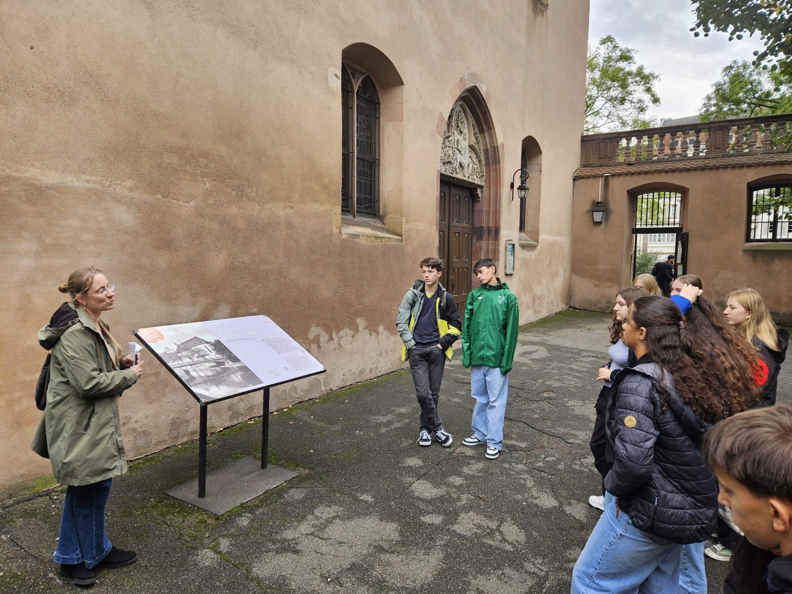 Eine Gruppe junger Menschen hört aufmerksam einer Frau mit Brille und grünem Mantel draußen zu.