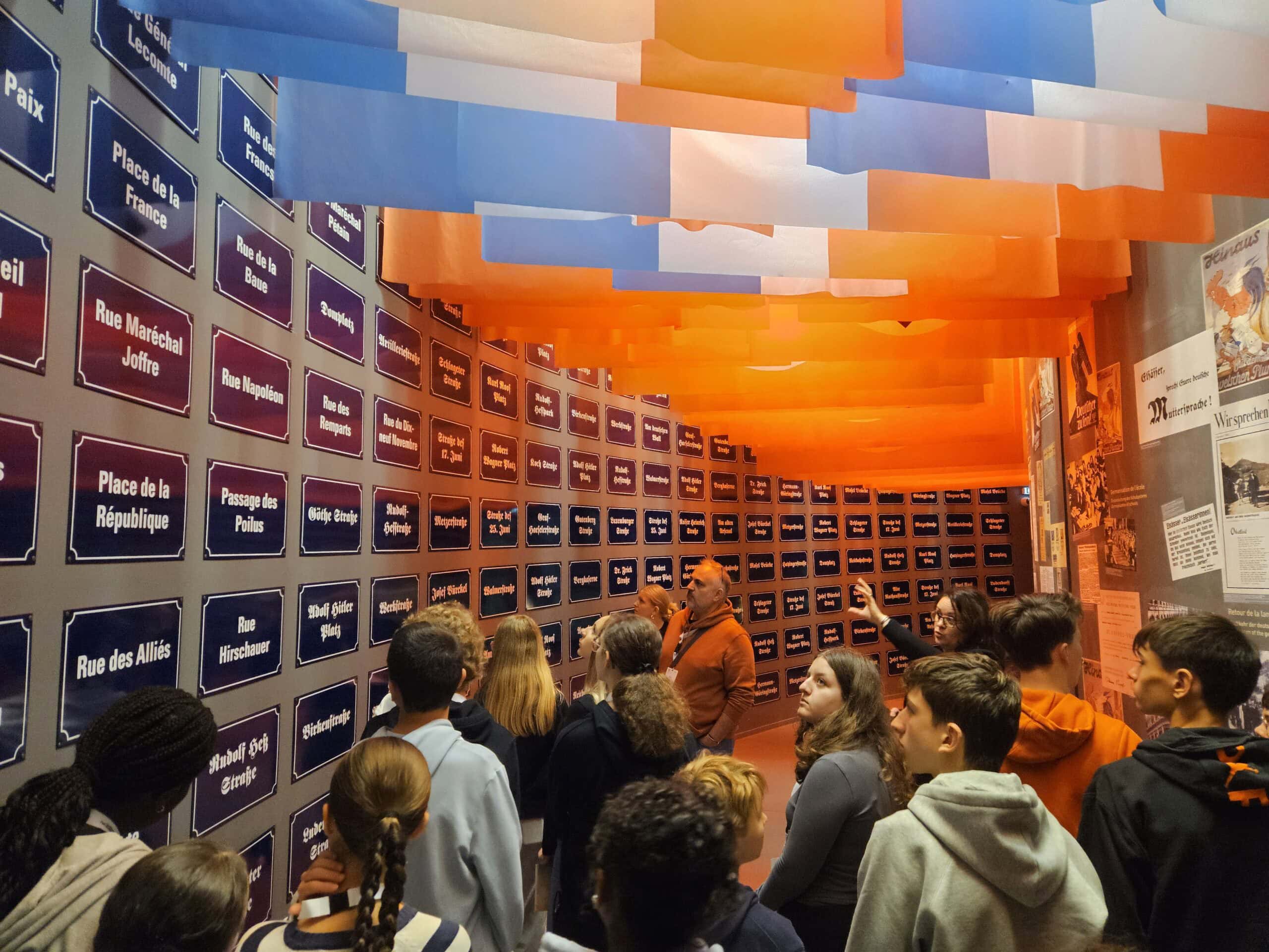 Besuchergruppe bei Ausstellung mit farbigen Flaggen, Person erklärt Schilder mit aufmerksamen Zuhörern.