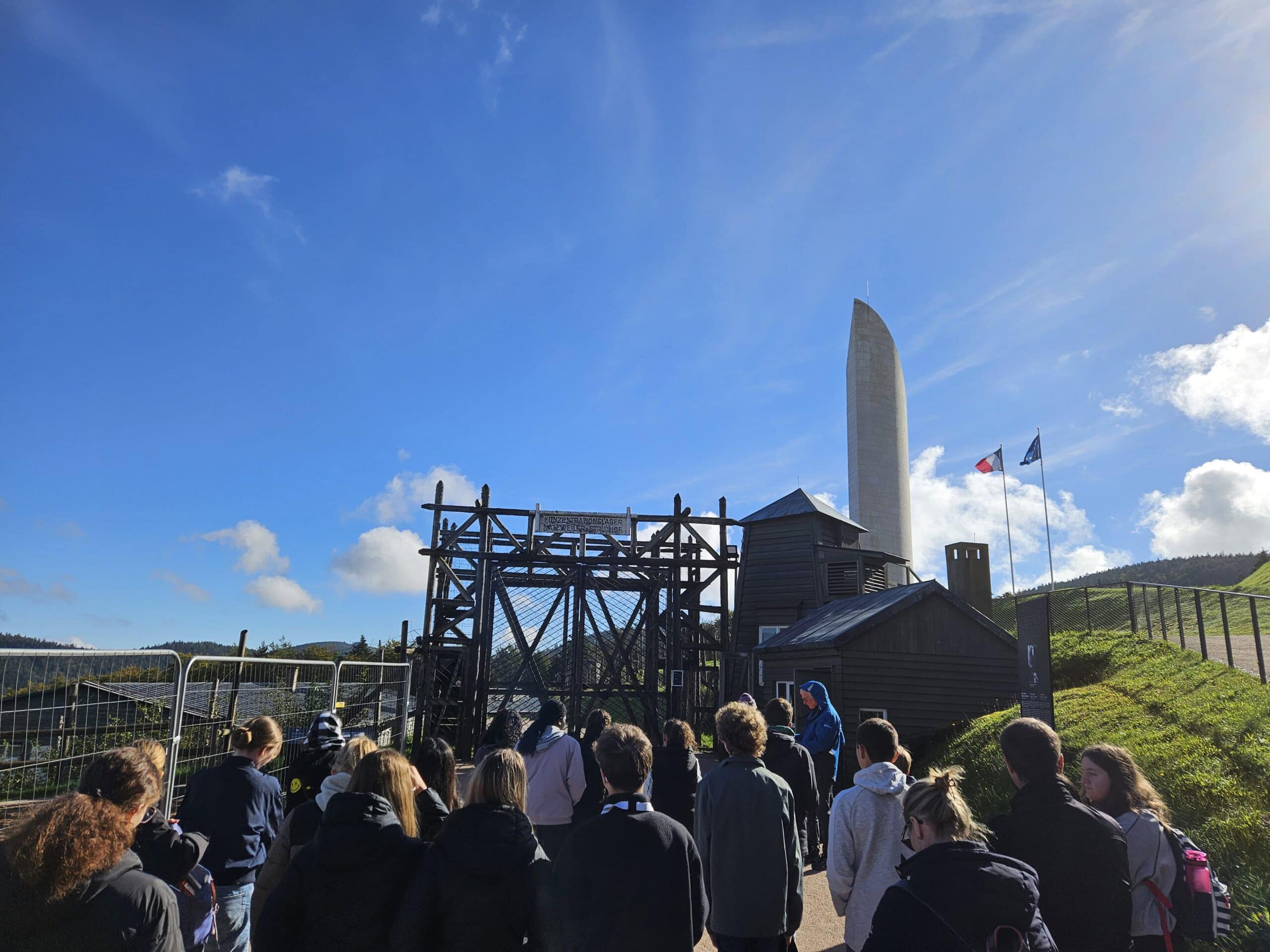 Gruppe von Menschen an einem Denkmal bei blauem Himmel, mit Fahnen und hölzernem Gebäude.