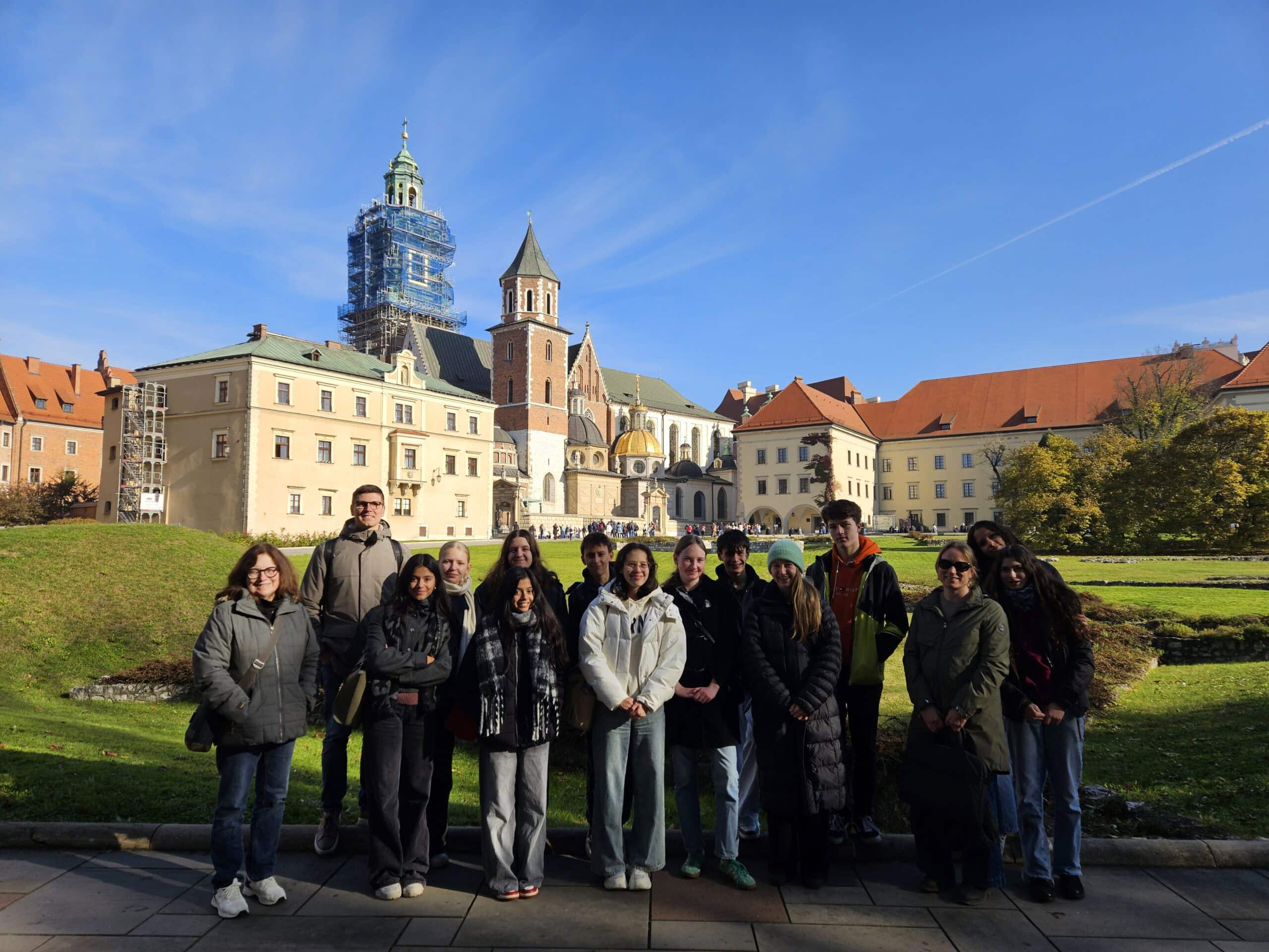 Gruppe von Menschen lächeln vor einem historischen Gebäude; blauer Himmel, sonnig.