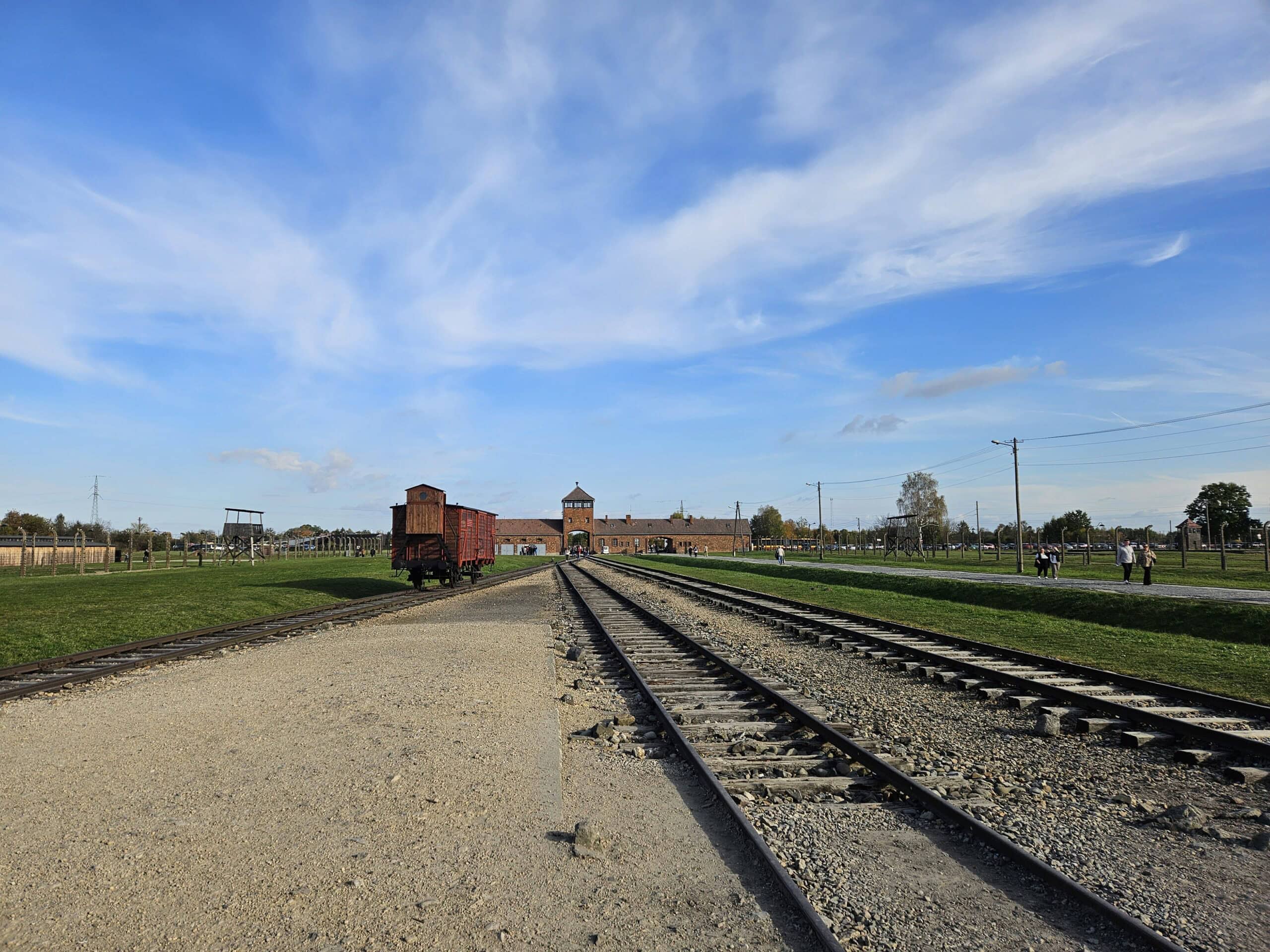 Weite Landschaft mit Bahngleisen, rotem Gebäude, blauem Himmel und vereinzelten Personen in der Ferne.