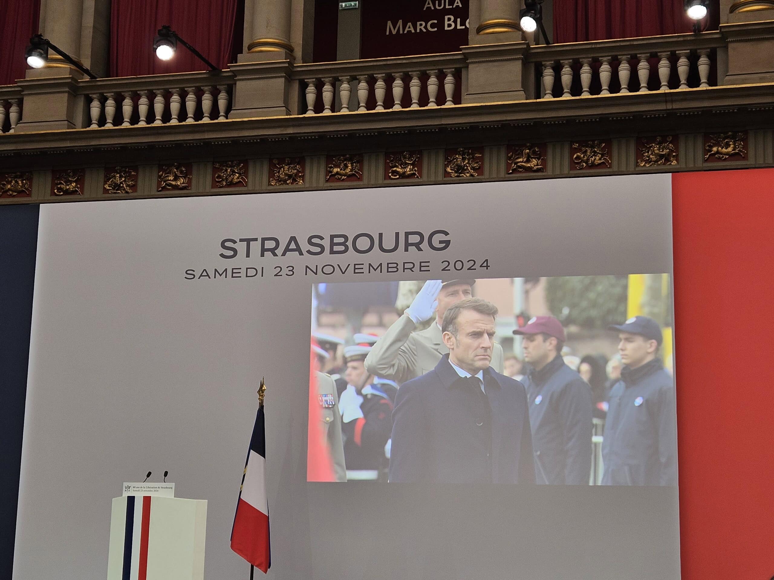 Ein Herr in einem dunklen Mantel, ernster Ausdruck, steht vor einem Bild mit einer Fahne.