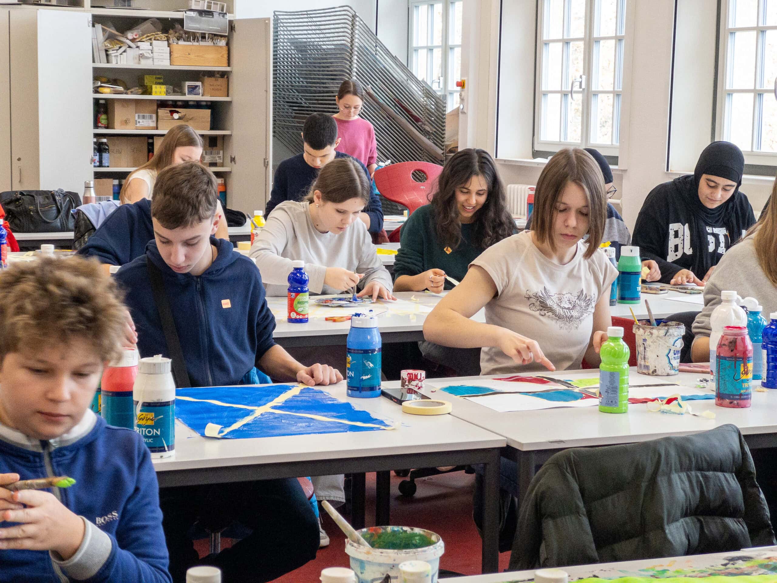 Eine Gruppe Jugendlicher malt konzentriert in einem Klassenzimmer voller bunter Farben.