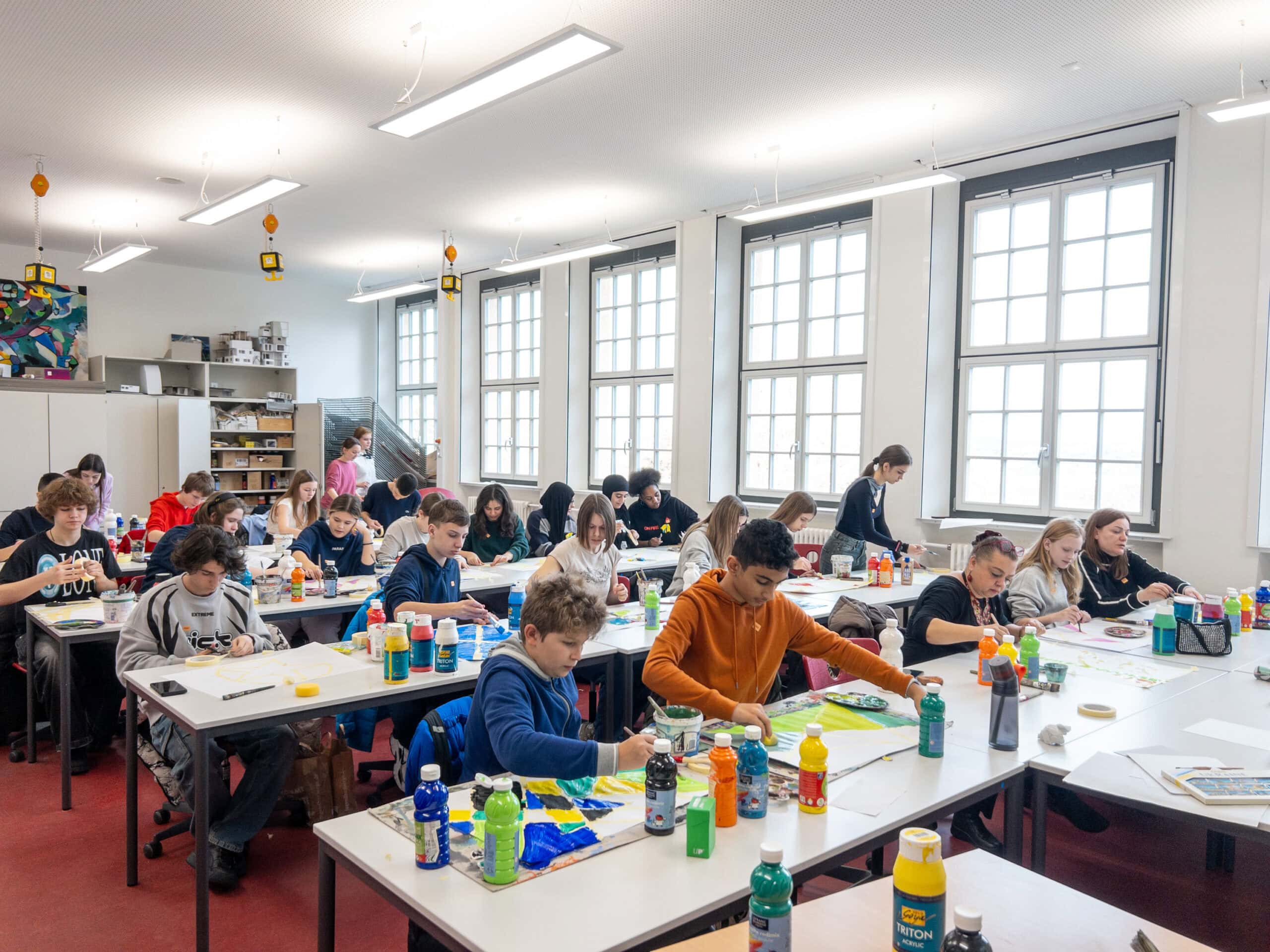 Kunstunterricht mit Schülern, die konzentriert an Tischen malen; helles Klassenzimmer, bunte Farben.