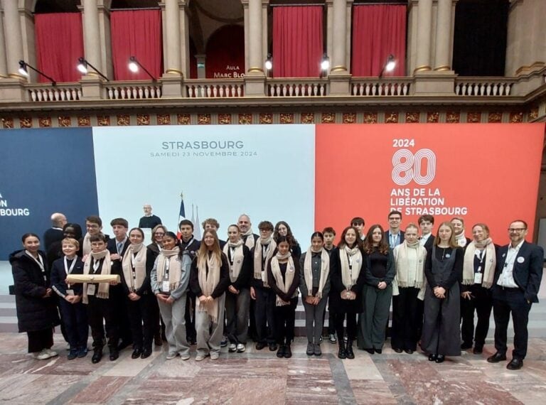 Eine Gruppe Jugendlicher und Erwachsener posiert lächelnd vor einer zweifarbigen Wand in Strasbourg.