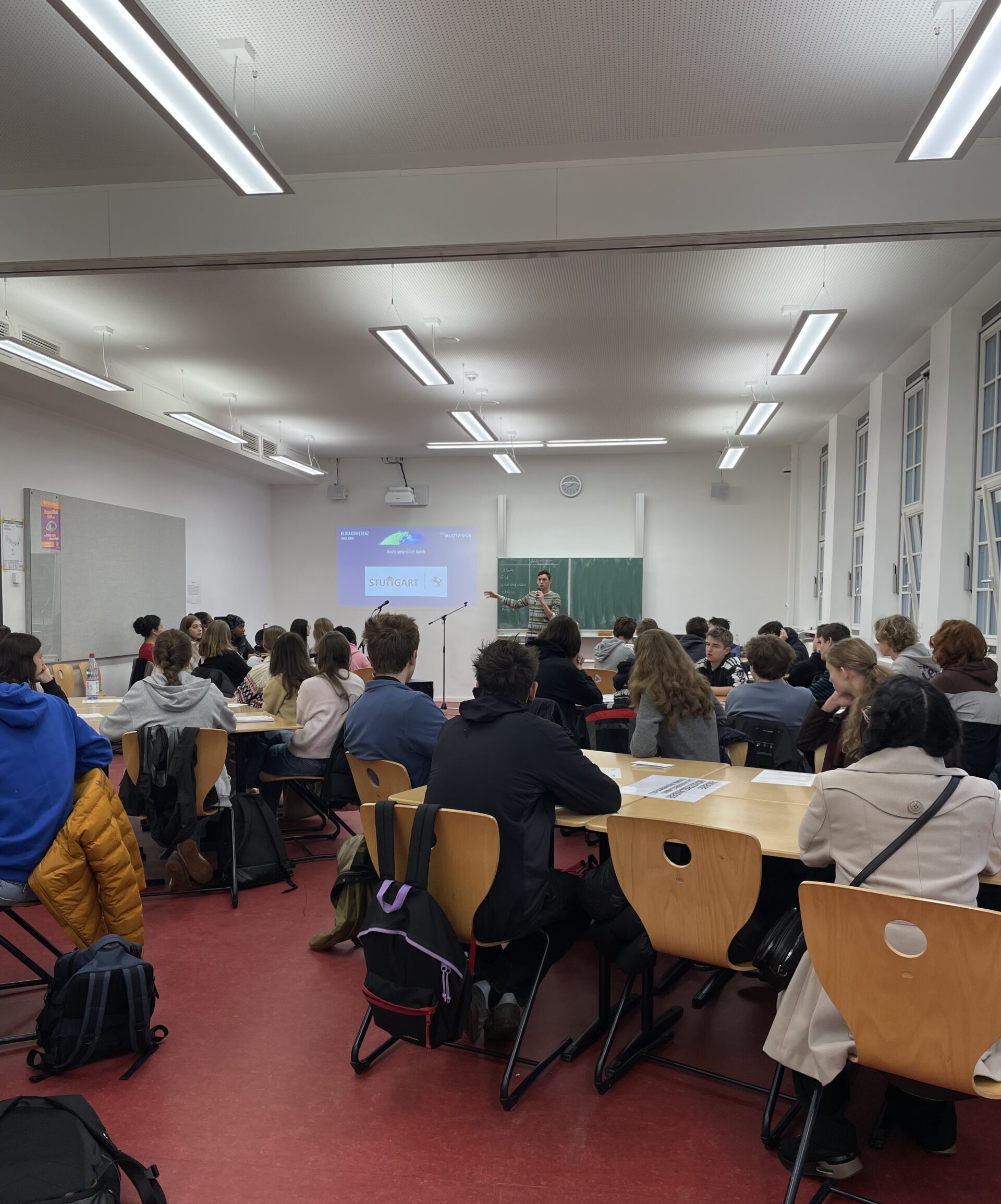 Eine Lehrkraft präsentiert in der Schule. Schüler hören aufmerksam zu. Helle Beleuchtung, rote Böden.