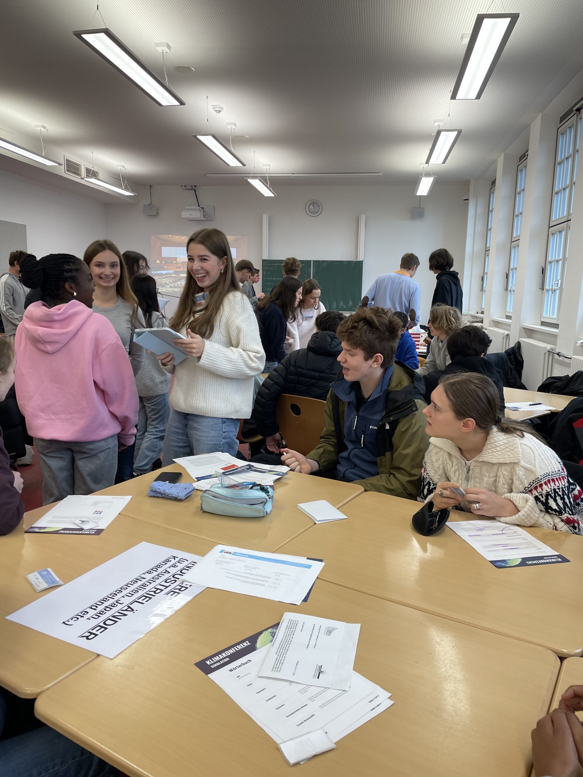 Junge Menschen lachen und unterhalten sich in einem Klassenzimmer mit hölzernen Tischen und Papieren.