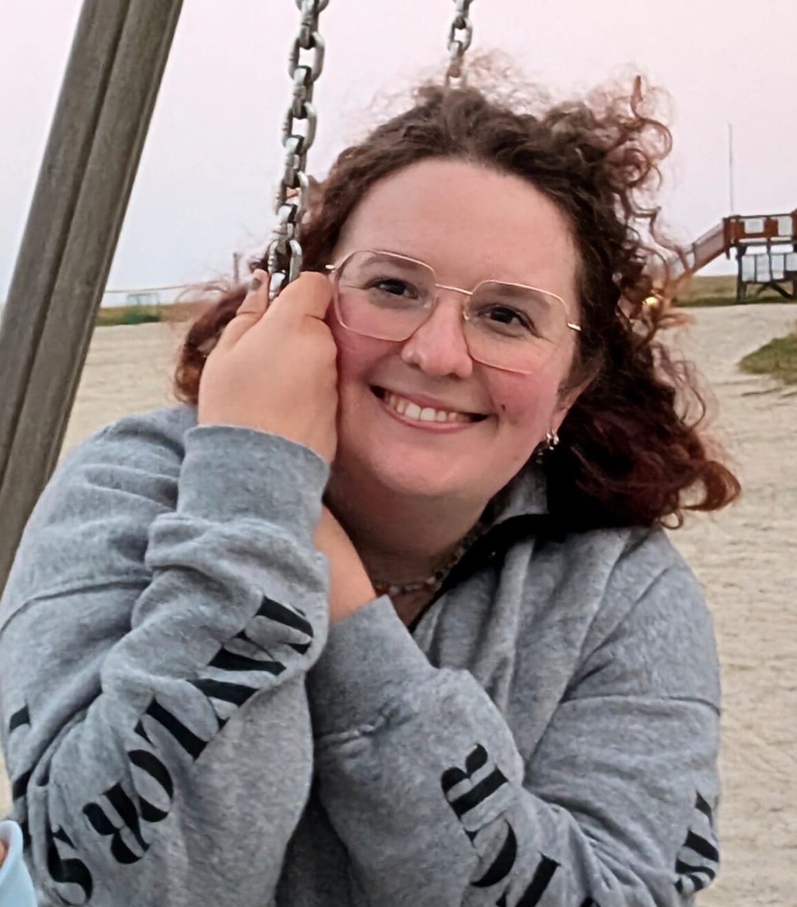 Person mit Brille lächelt auf einer Schaukel am Strand, trägt grauen Hoodie.