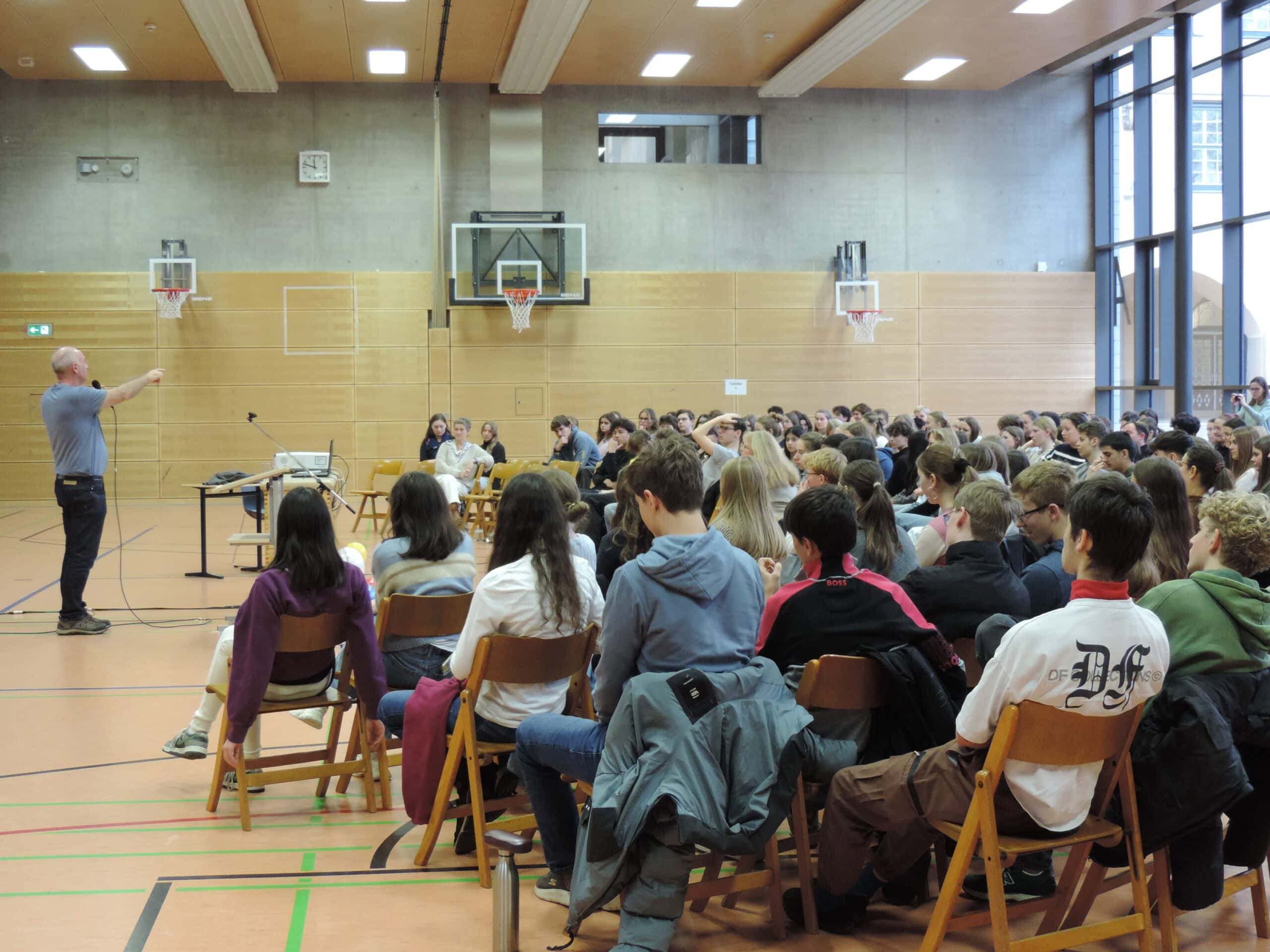 Eine Person hält in der Turnhalle vor sitzendem Publikum einen Vortrag, alle aufmerksam lauschend.