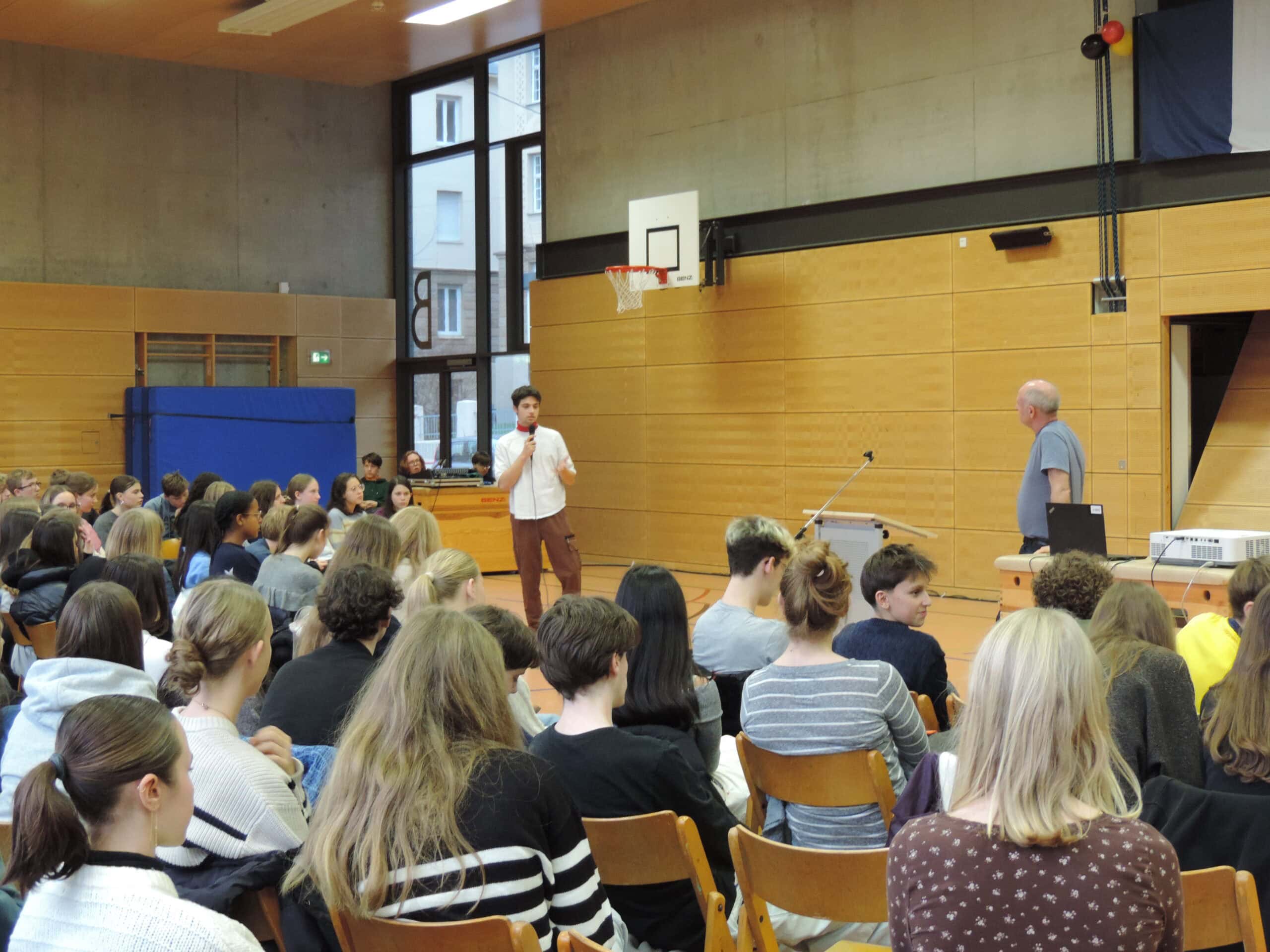 Ein Mann spricht vor Schülergruppen in einer Turnhalle. Die Wände sind aus hellem Holz.