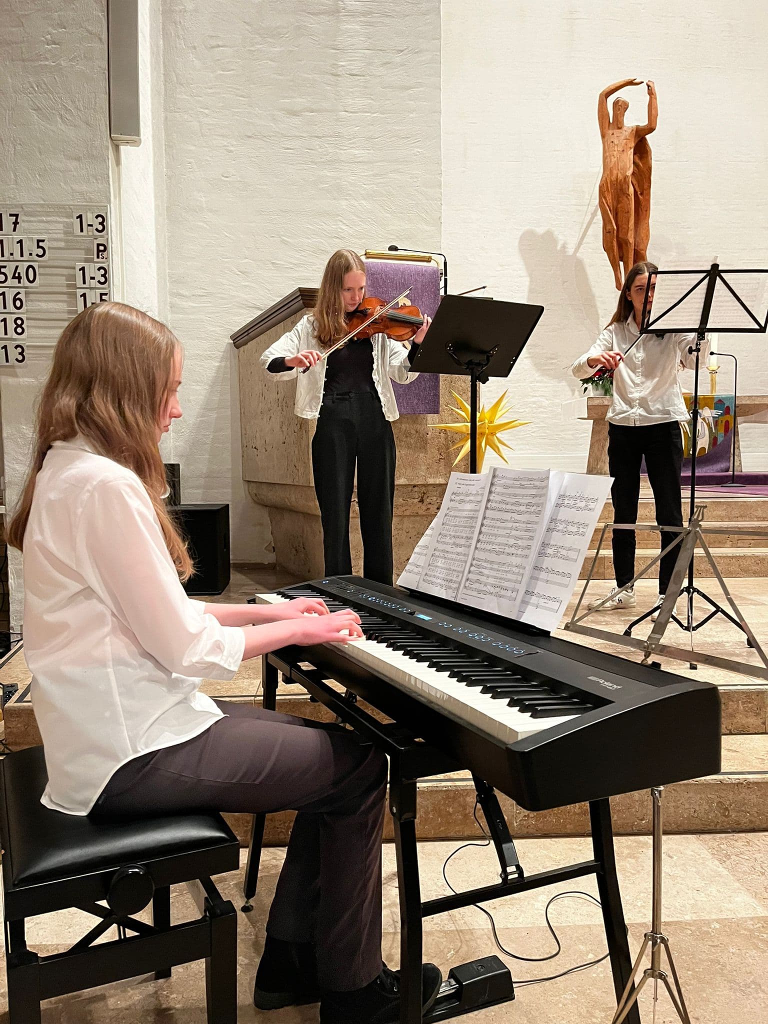 Drei junge Frauen musizieren in heller Kirche, spielen Klavier und Geige, konzentrierte Ausdrücke.