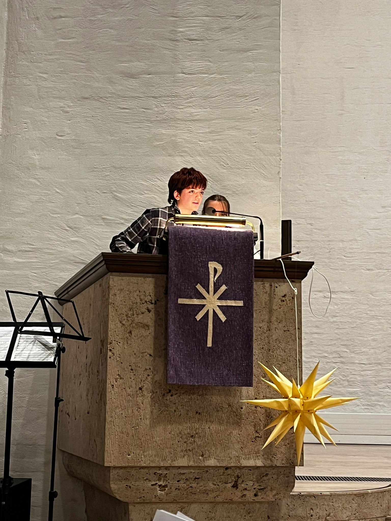 Person am Rednerpult mit ernstem Gesichtsausdruck, lila Banner, gelber Stern im Vordergrund.