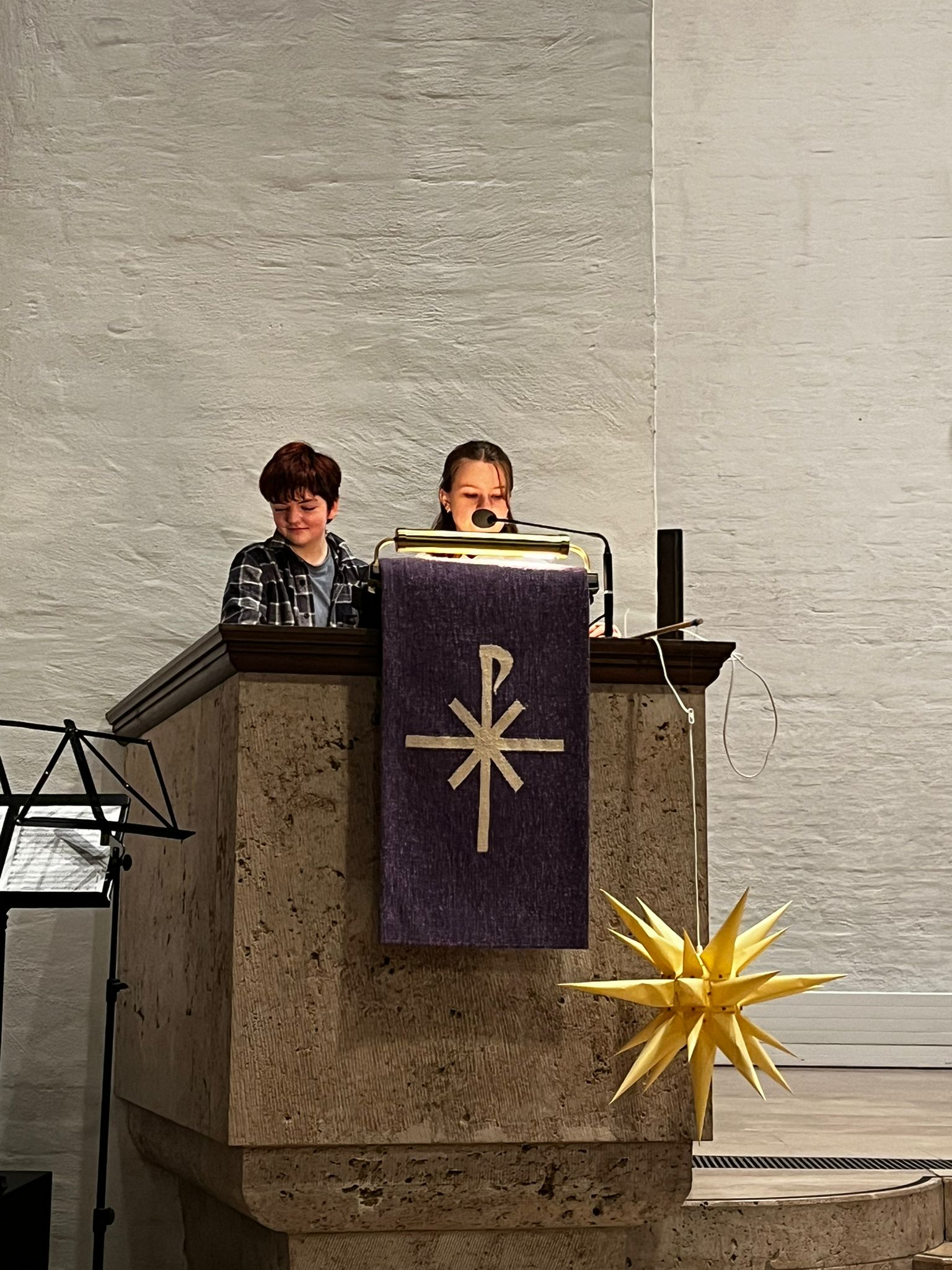 Zwei Jugendliche am Rednerpult in der Kirche, lächelnd, mit violettem Banner und gelbem Stern.