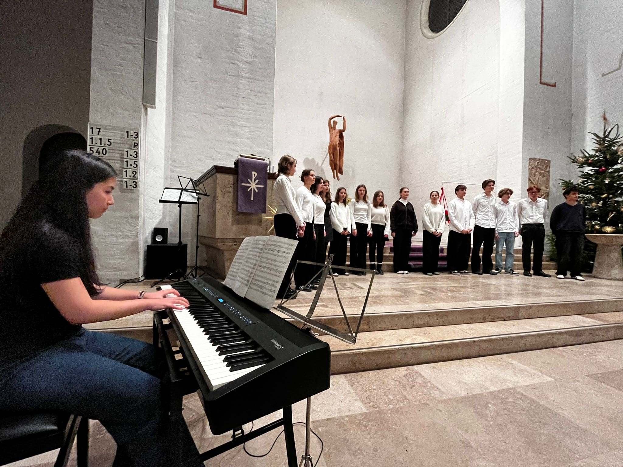 Frau spielt Klavier, Chor singt lächelnd in Kirche mit weißer Wand, Weihnachtsbaum rechts.