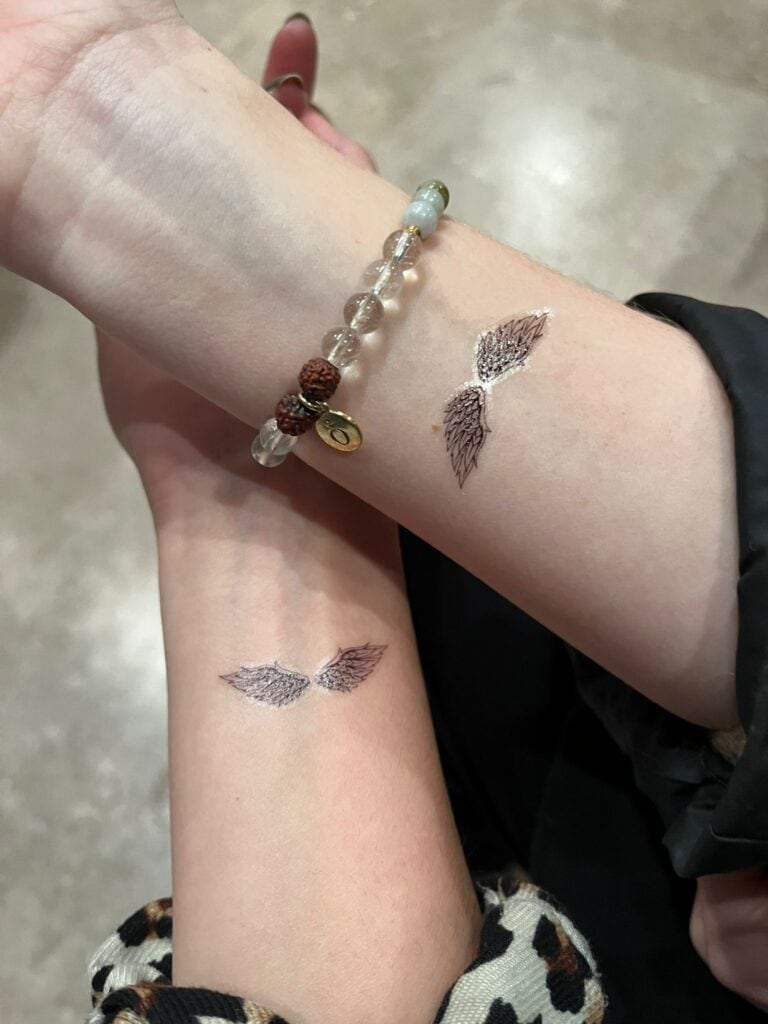 Zwei Unterarme mit identischen Flügel-Tattoos. Ein Arm trägt ein Armband mit Glasperlen.