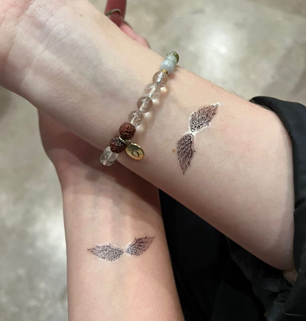 Zwei Unterarme mit identischen Flügel-Tattoos. Ein Arm trägt ein Armband mit Glasperlen.