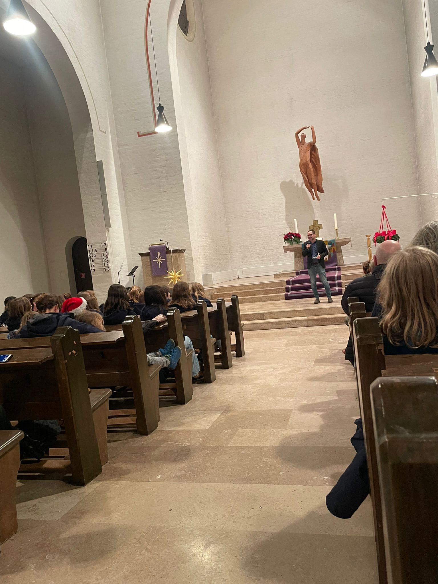 Eine Person spricht in einer Kirche, lächelnd. Holzbänke, weiße Wände, rote Kerzen, Statue oben.