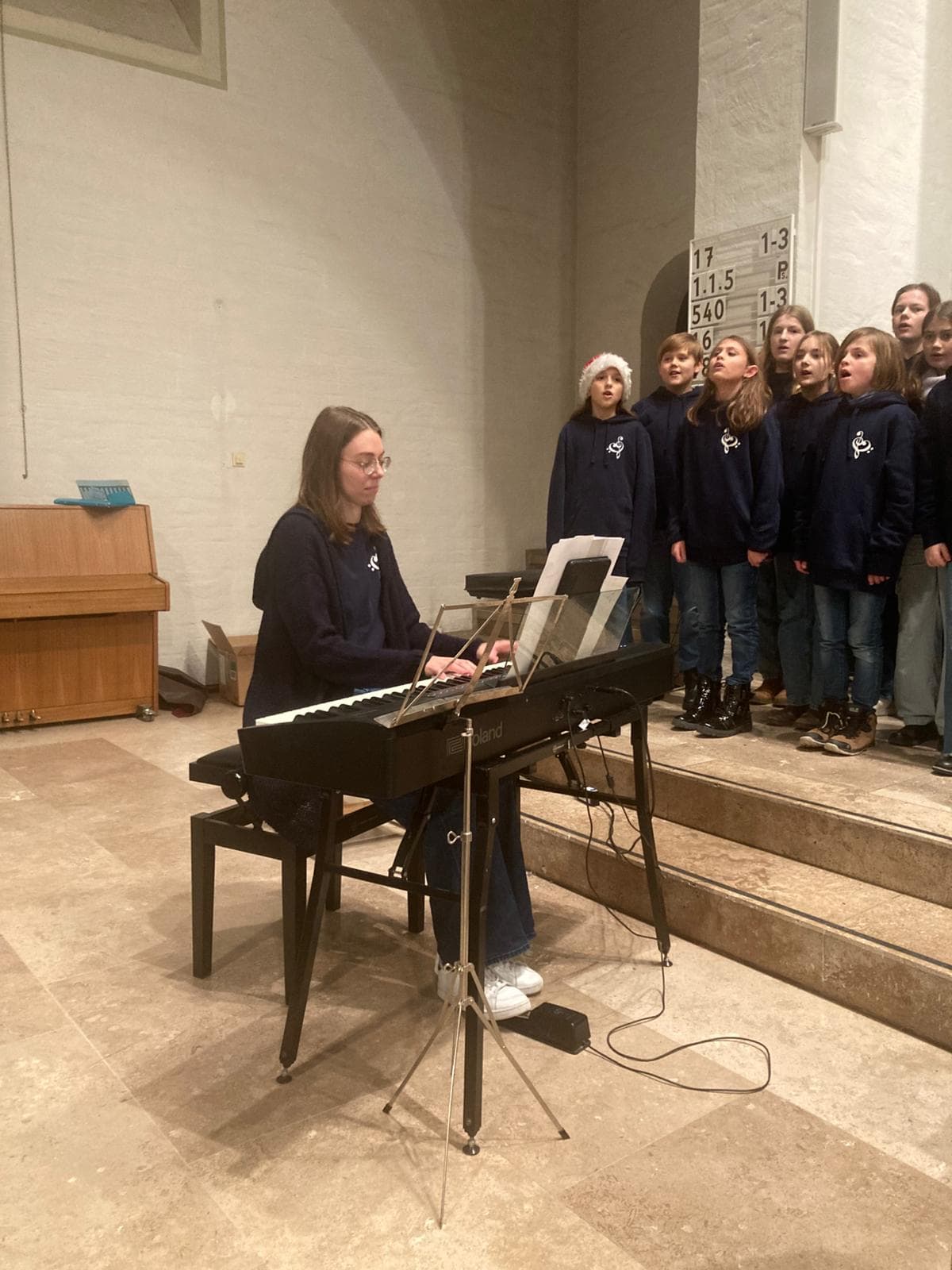 Eine Frau spielt fröhlich Klavier, Kinderchor in blauen Pullovern rechts auf Stufen.