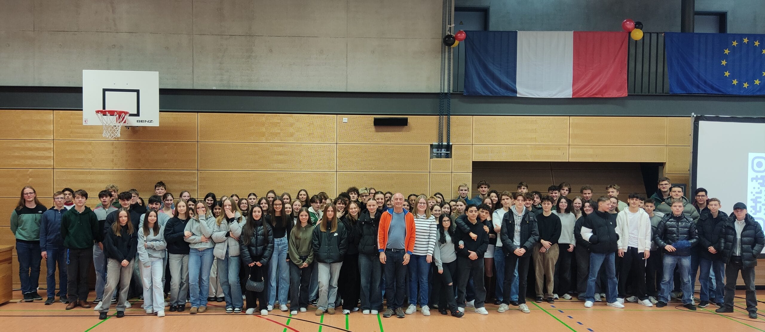 Eine große Gruppe von Schülern posiert lächelnd in einer Turnhalle mit Basketballkorb.