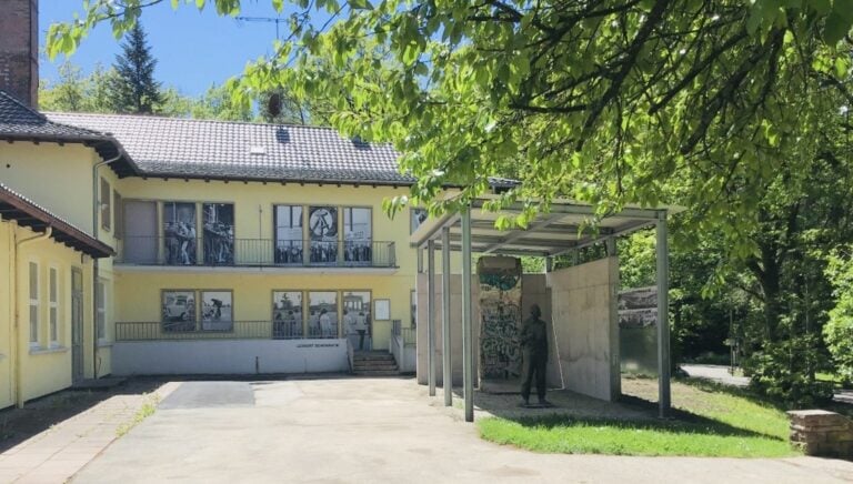 Gelbes Gebäude mit Wandkunst und schattiger Terrasse, umgeben von grünen Bäumen bei Sonnenschein.