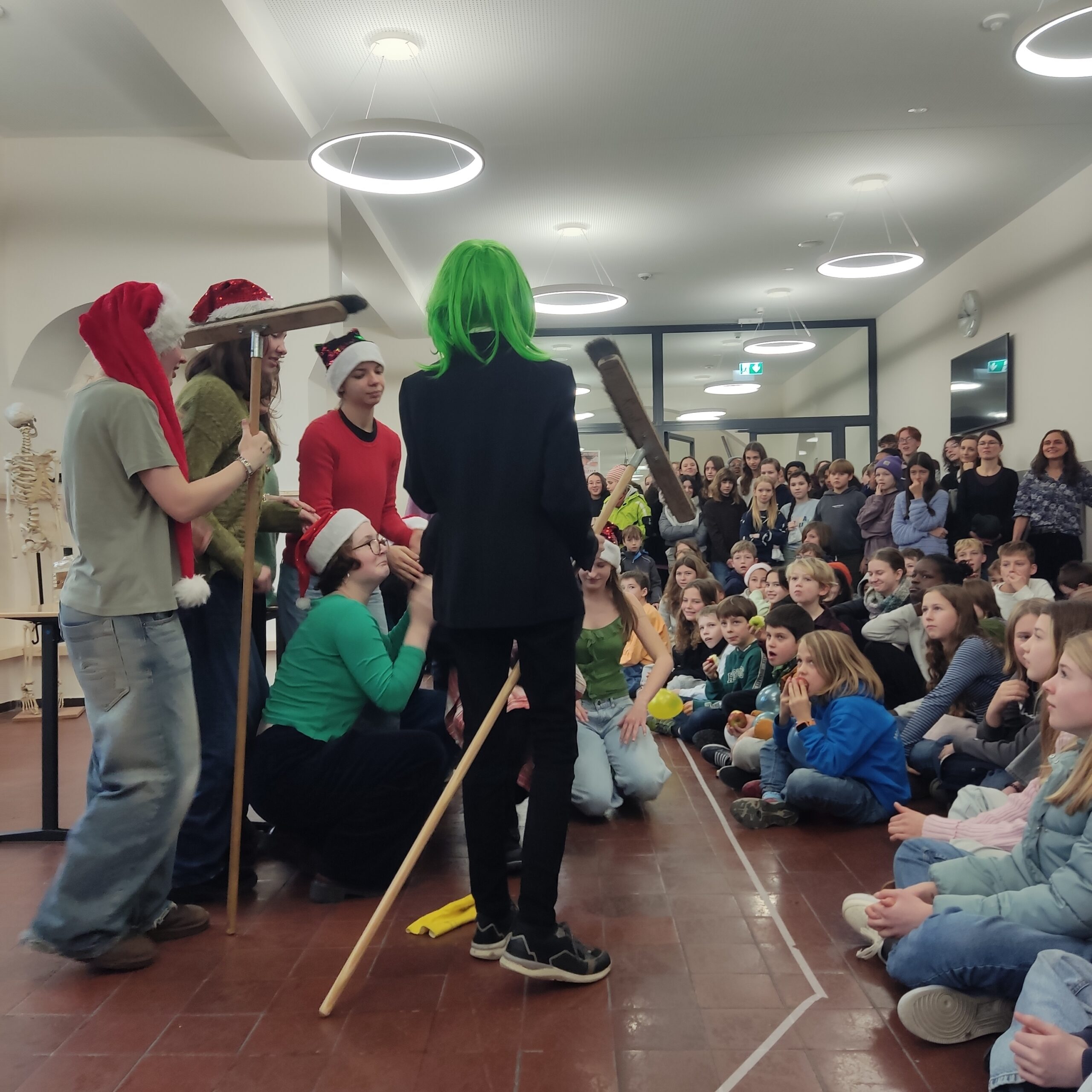 Eine Personengruppe mit Weihnachtsmützen, lachend, steht vor einem sitzenden Publikum; bunte Haare.