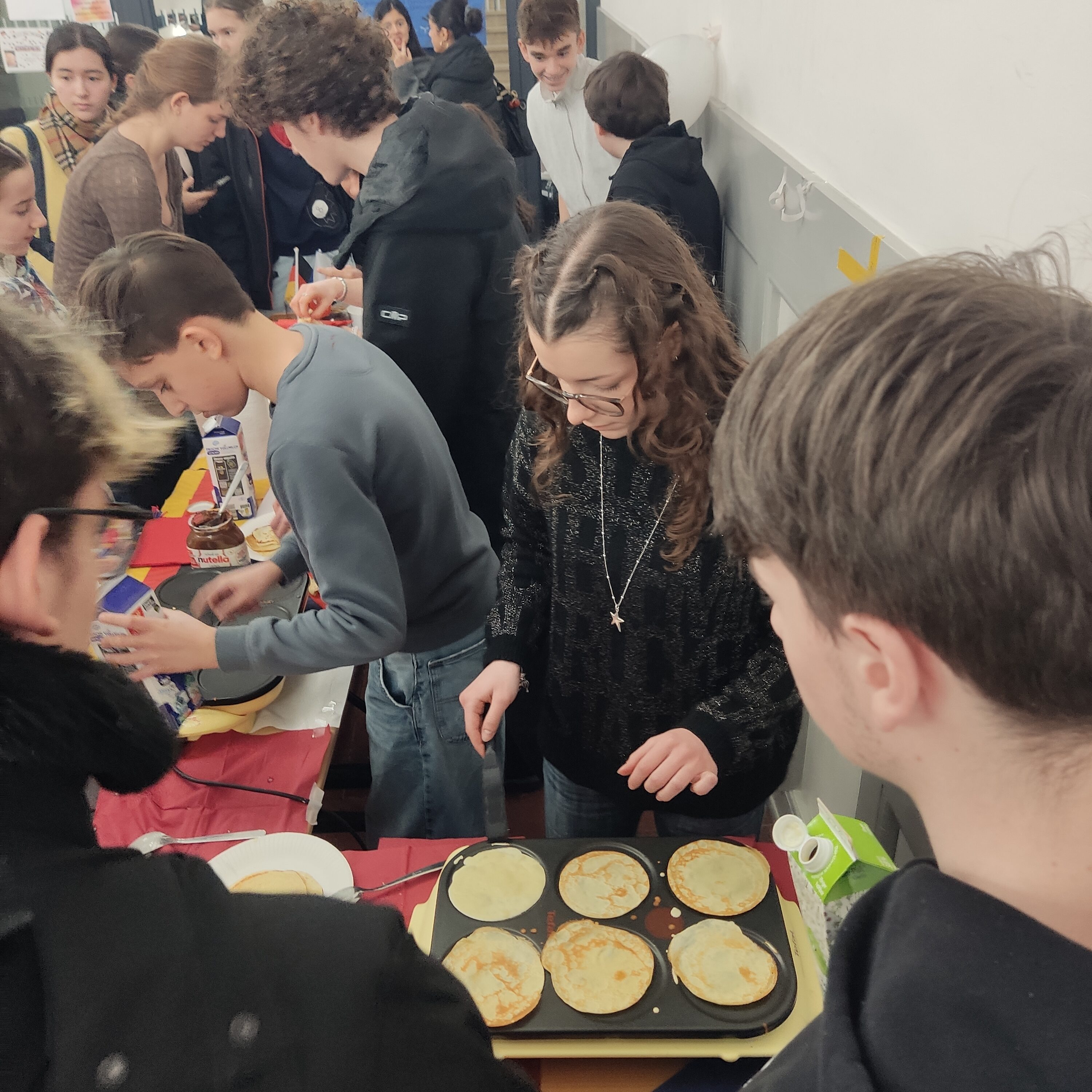 Eine Frau mit lockigem Haar macht Pfannkuchen auf einem Tisch inmitten einer Gruppe junger Leute.