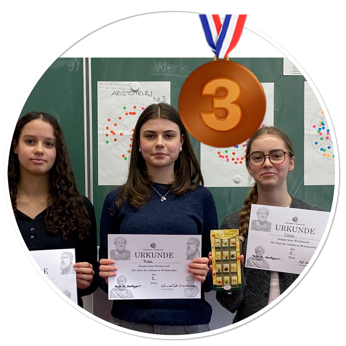 Drei junge Frauen lächeln mit Urkunden vor einer Tafel; eine trägt eine Medaille.