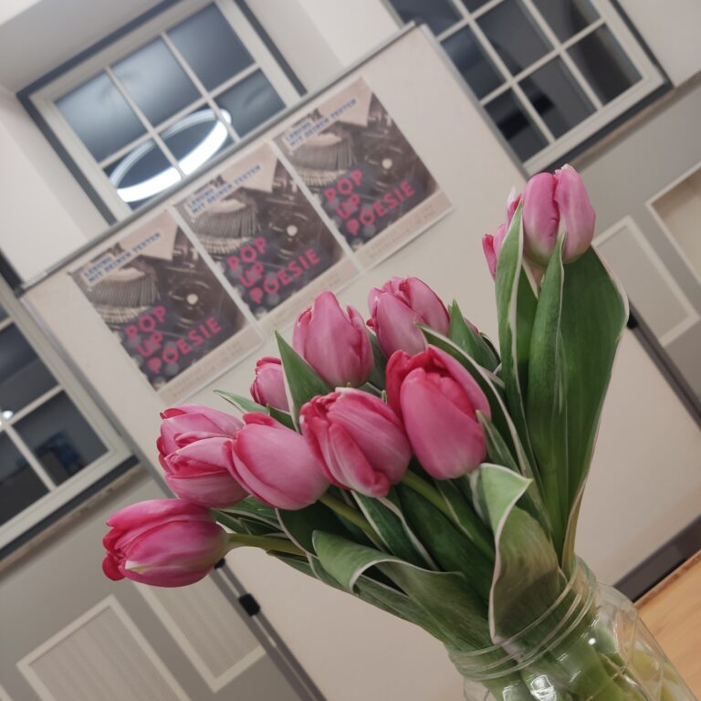 Ein Strauß pinker Tulpen in einer Vase vor einem Fenster mit Postern im Hintergrund.