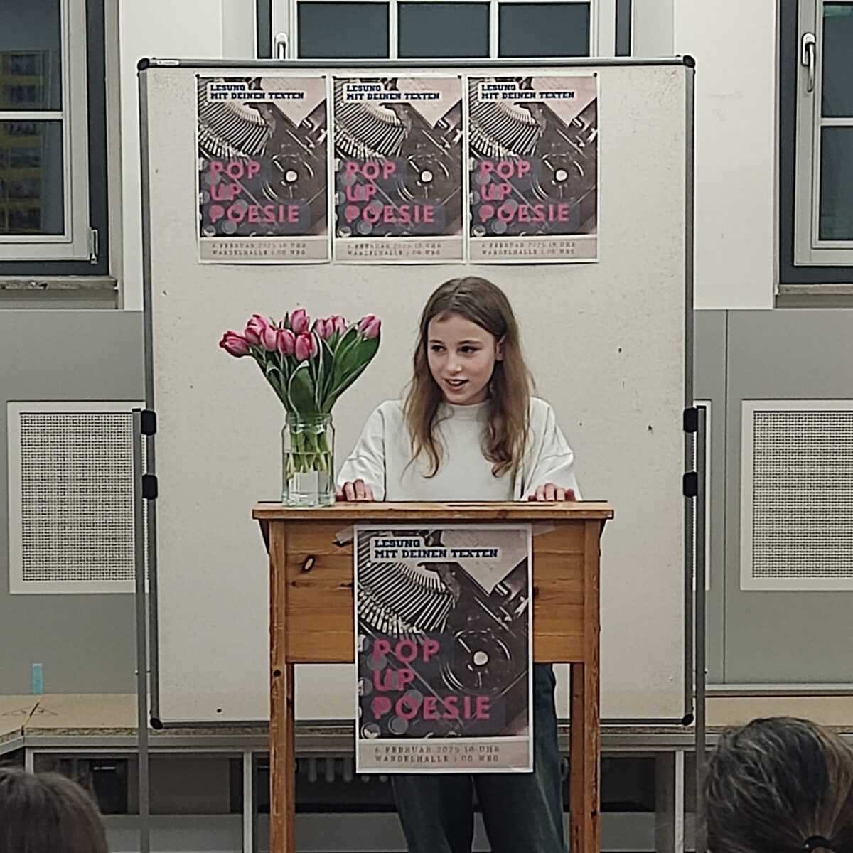 Frau hält Rede, lächelt, weiße Bluse, Tulpen auf Tisch, Poster im Hintergrund.